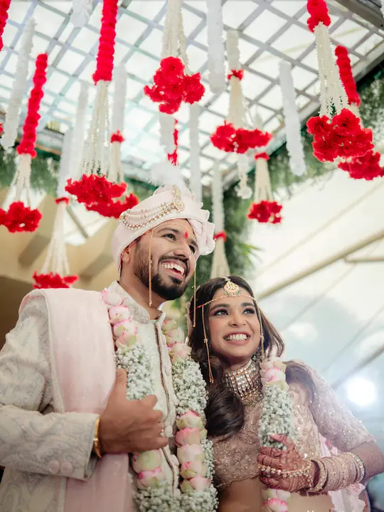 Rahul and Arundhati smiling together beneath hanging red flowers at their wedding.